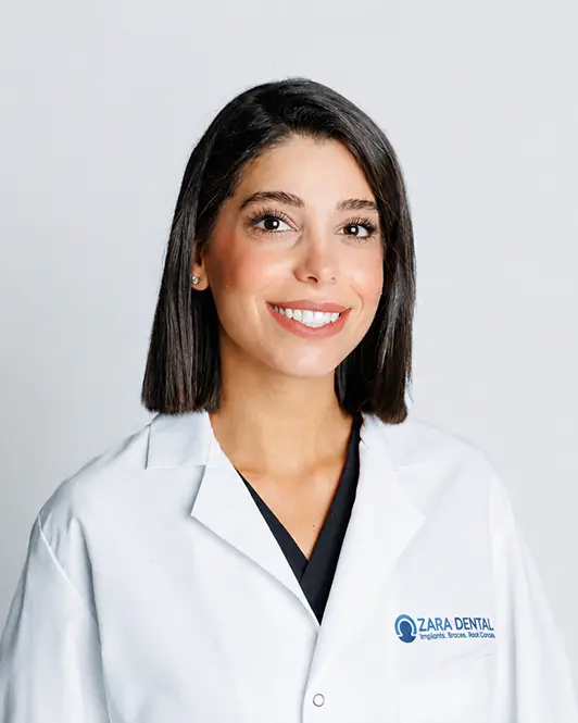 Smiling woman in a white dental coat with the Zara Dental logo, posing for a portrait.